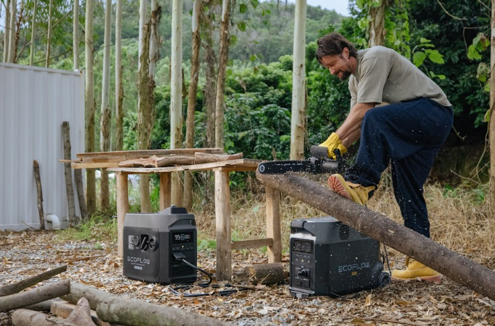 Person wearing safety goggles operates a circular wood saw outdoors, powered by a compact dual-fuel generator (LPG/petrol) positioned nearby—demonstrating reliable emergency power in action.